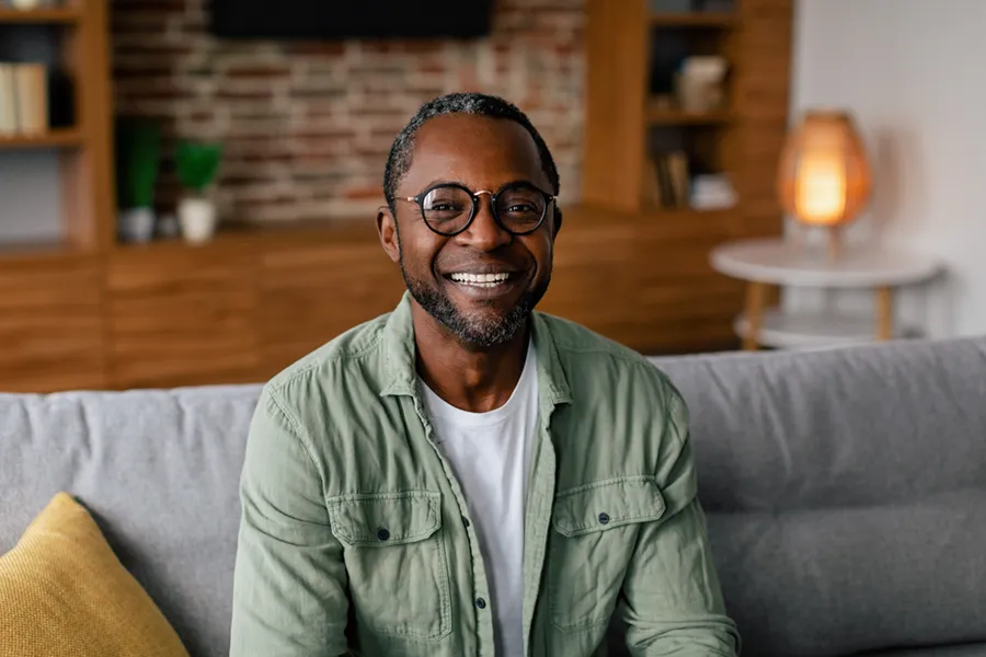 Andropause-Doctor A middle aged black man in a green button up shirt sits on his couch smiling, happy with his treatment for andropause and low testosterone from Sandra McDonald, NP in Southwestern Ontario.