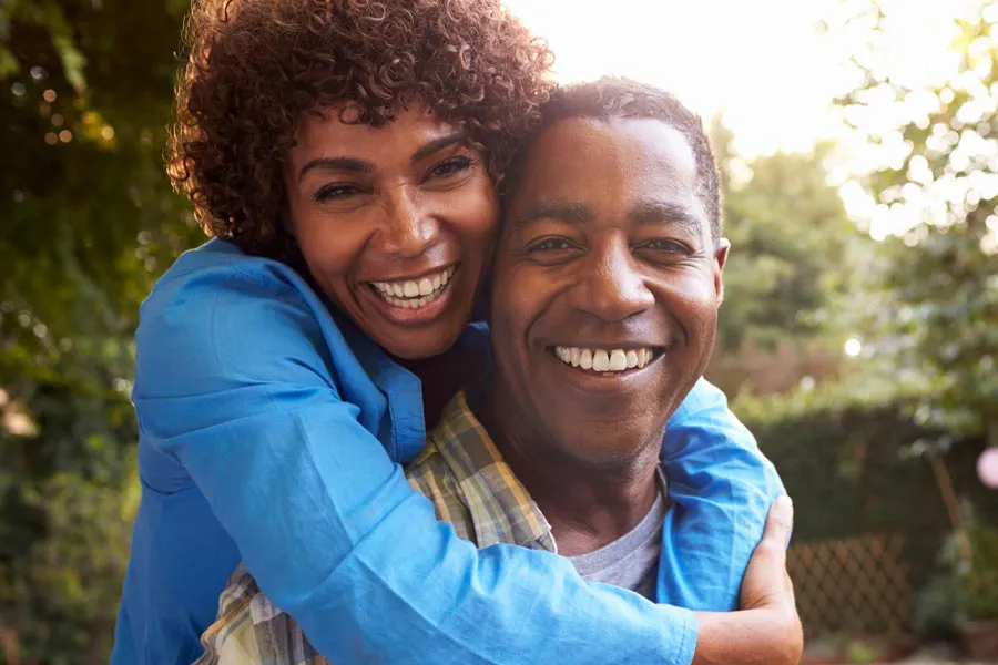 Autoimmune-Doctor A middle-aged African American couple outside, the woman embracing the man from behind pressing her cheek to his, both are smiling due to successful Autoimmune Disorder treatment from Sandra McDonald, NP in Southwestern Ontario.
