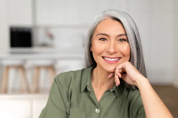 Menopause-Clinic An asian woman with long gray hair and a green shirt sitting and smiling on her couch, happy with her menopause care from [NAME} in Southwestern Ontario.