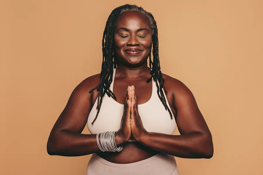 Obesity-Clinic An older African American woman confidently posing against a warm background smiling with hands in a prayer pose. Sandra McDonald, NP treats obesity using functional medicine in Southwestern Ontario.