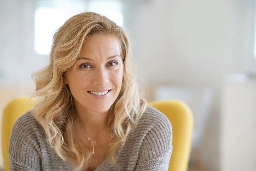 Perimenopause-Doctor A blonde woman wearing a a gray sweater sits inside in a yellow chair, smiling and happy with her perimenopause treatment from Sandra McDonald, NP in Southwestern Ontario.